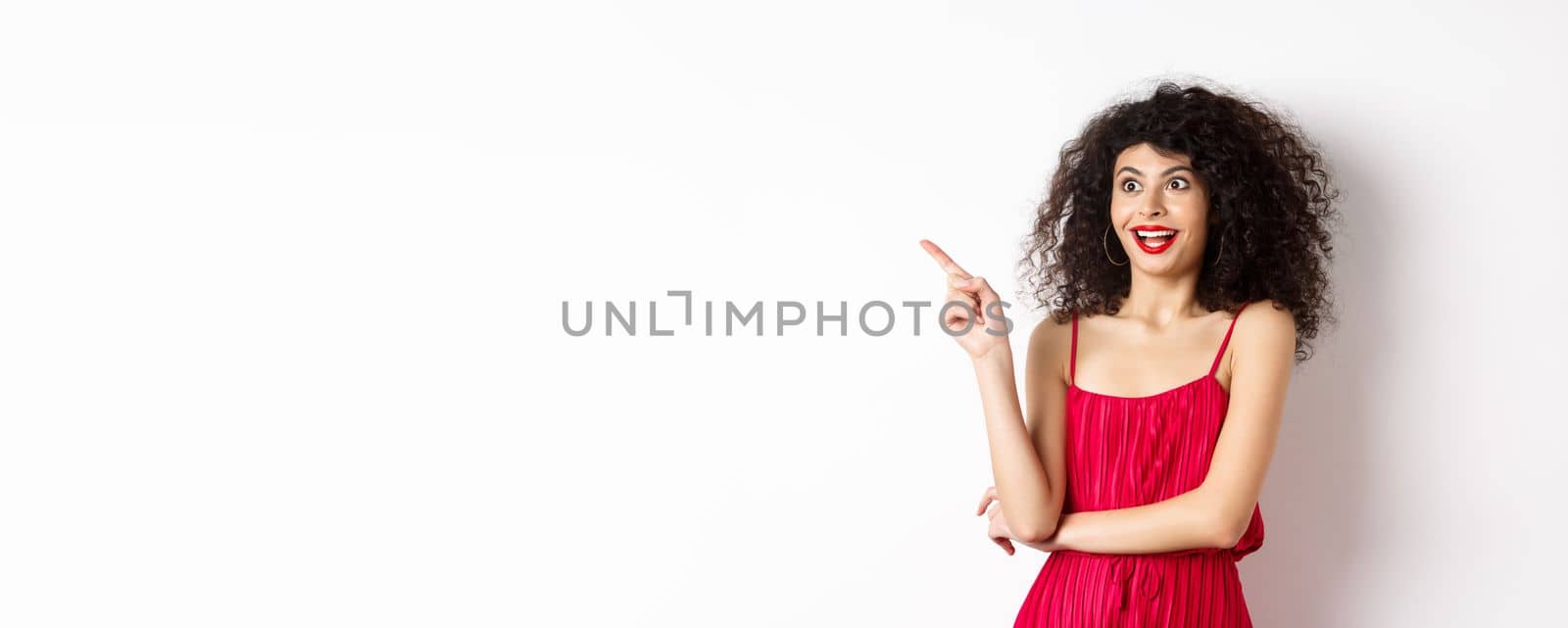 Surprised female model with curly hair and makeup, pointing finger and looking left with amused smile, standing in red dress over white background.