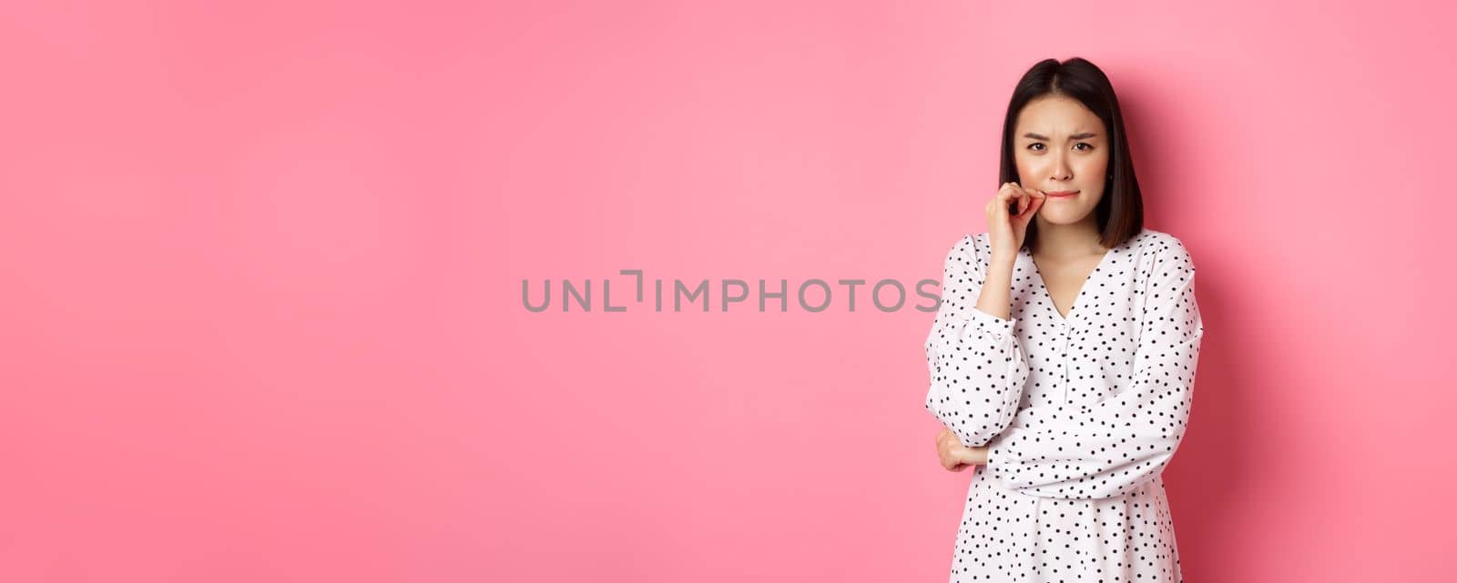 Brunette asian woman in dress looking displeased, frowning and zipping mouth, seal lips with promise, standing over pink background by Benzoix