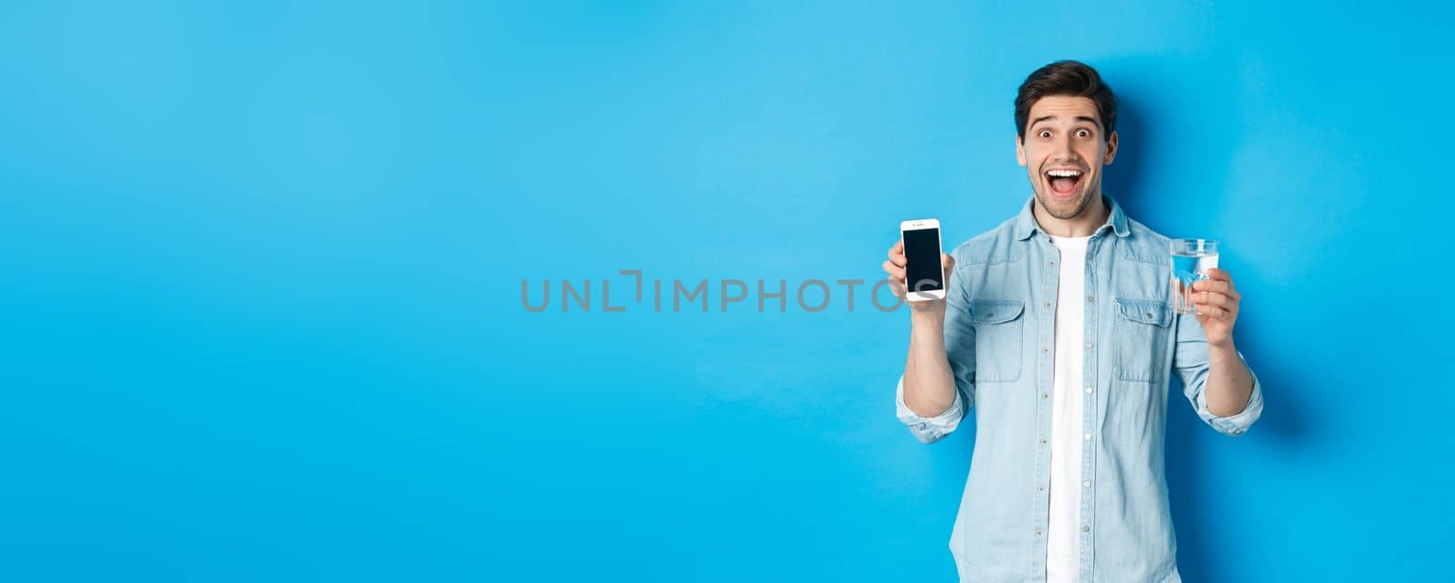 Happy man showing glass of water and mobile screen, recommending smartphone health app, standing over blue background by Benzoix