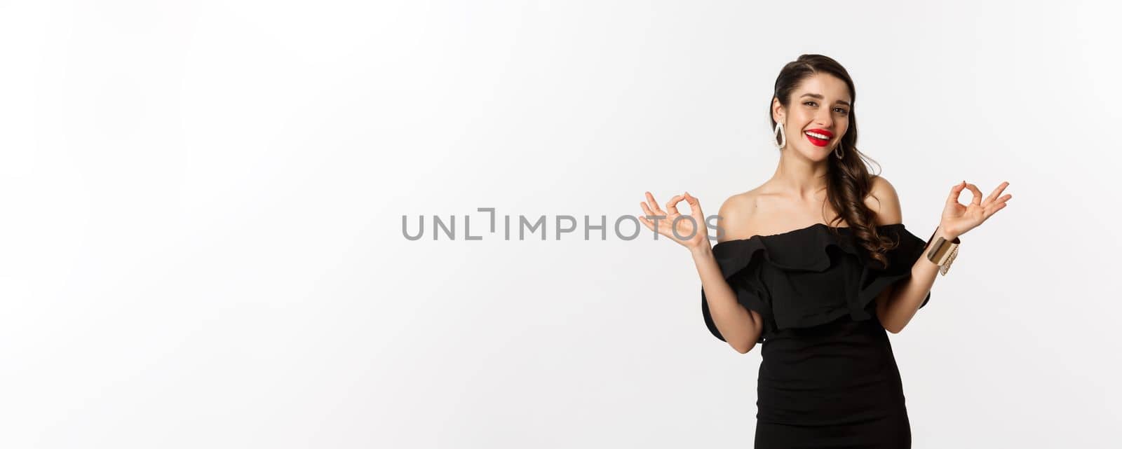 Fashion and beauty. Attractive brunette woman in black dress, showing okay signs and smiling satisfied, approve and recommend, standing over white background.