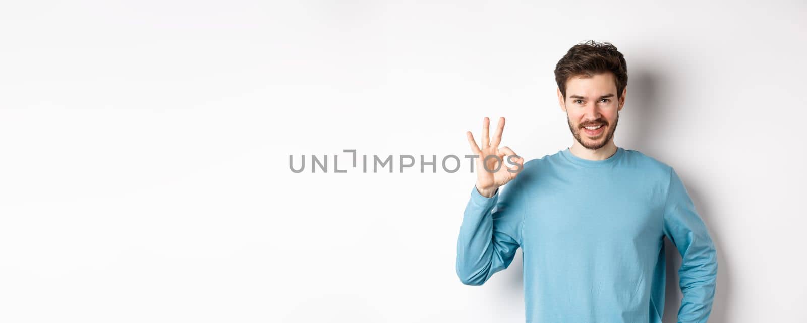 Okay. Handsome smiling man showing ok sign and looking confident, recommend or praise something good, standing over white background.