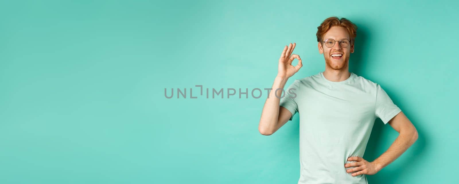 Happy young man with red hair and beard, wearing glasses and t-shirt, smiling satisfied and showing ok sign, say yes, approve and agree, standing over turquoise background by Benzoix