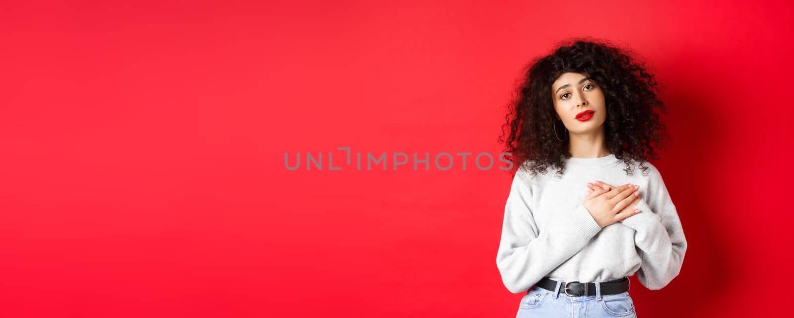 Beautiful woman with curly hair feeling touched and thankful, holding hands on heart and looking with affection at camera, red background.