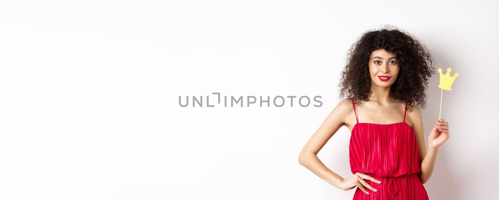 Stylish young woman in red dress, feeling confident and sassy, holding crown and smiling, standing over white background.