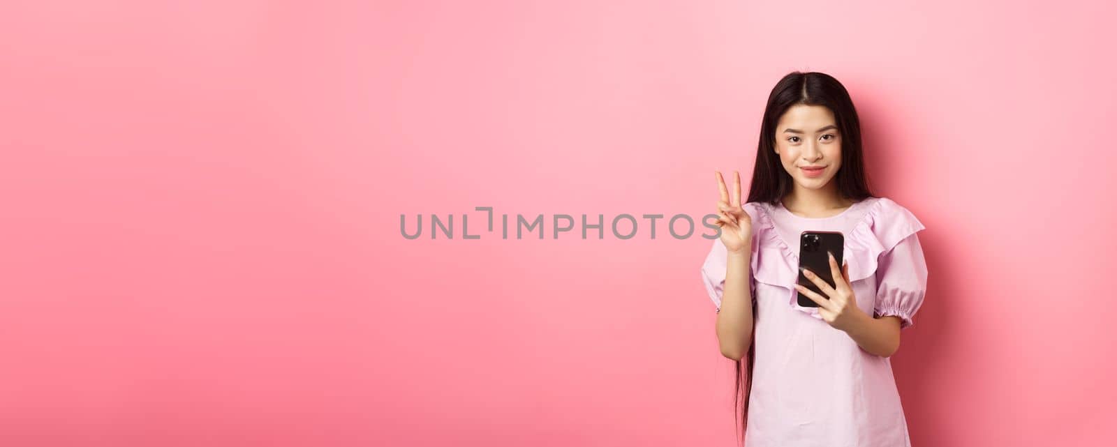 Online shopping. Stylish asian teen girl using smartphone, show peace sign and smiling pleased at camera, standing on pink background by Benzoix