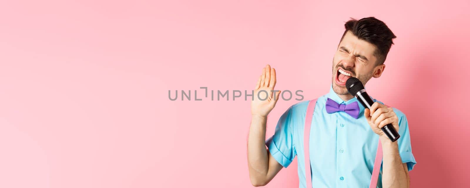 Party and festive events concept. Funny guy performs a song, singing karaoke in microphone with carefree face, standing in bow-tie and suspenders on pink background by Benzoix