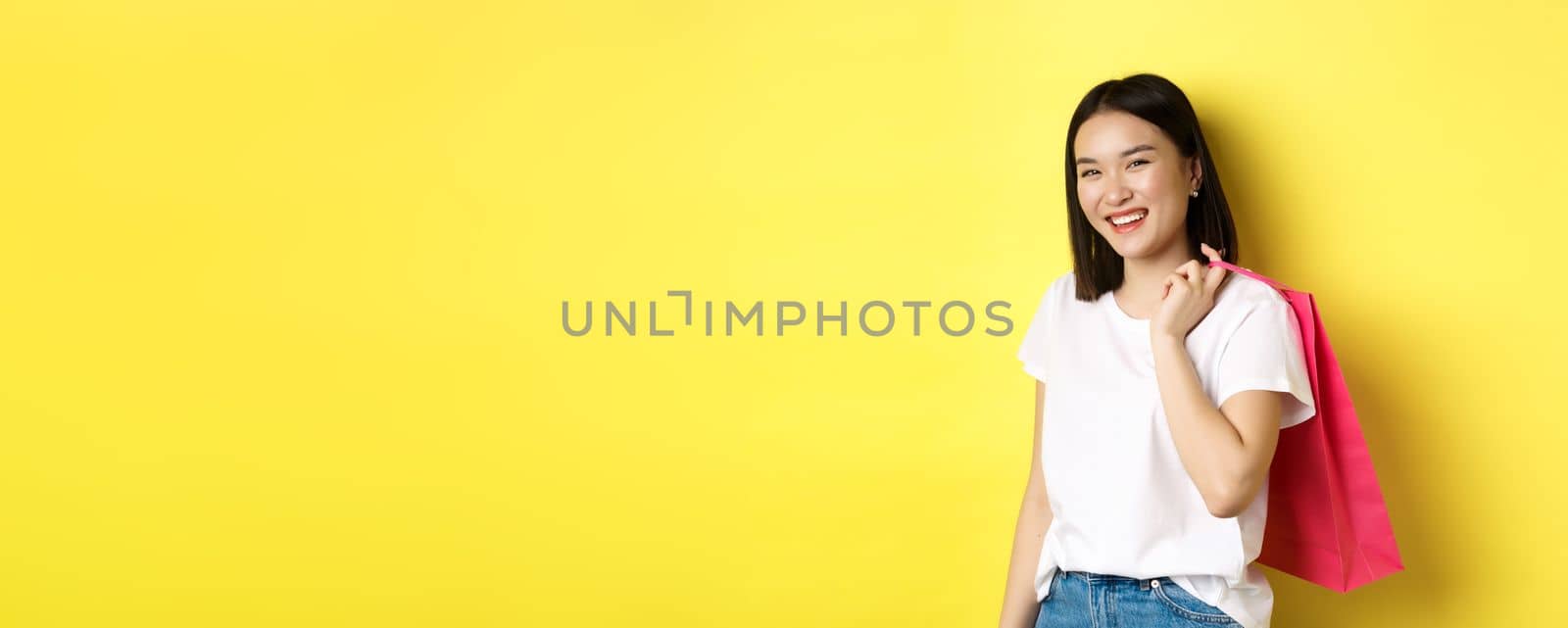 Happy asian woman holding shopping bag behind shoulder and laughing, standing over yellow background.