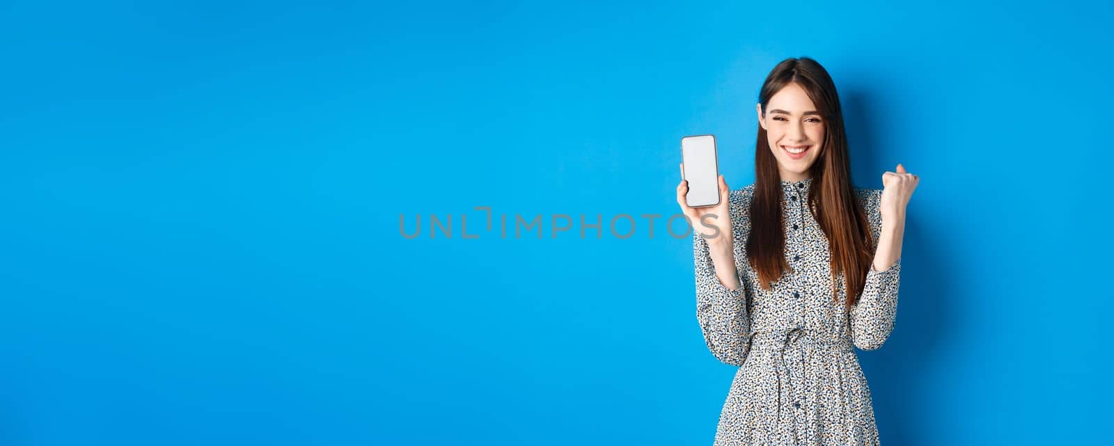 Cheerful pretty girl chanting, winning on mobile phone, showing empty smartphone screen and fist pump, smiling and celebrating, blue background by Benzoix