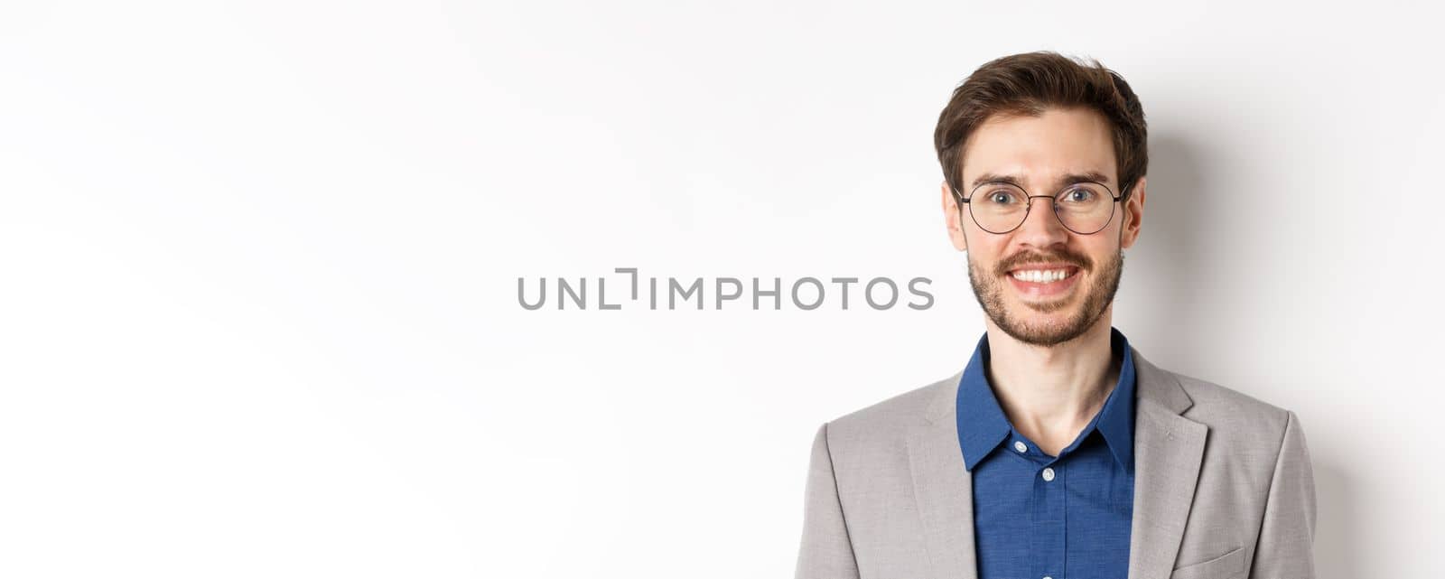 Close-up of handsome young office worker in glasses and suit smiling at camera, looking happy on white background by Benzoix