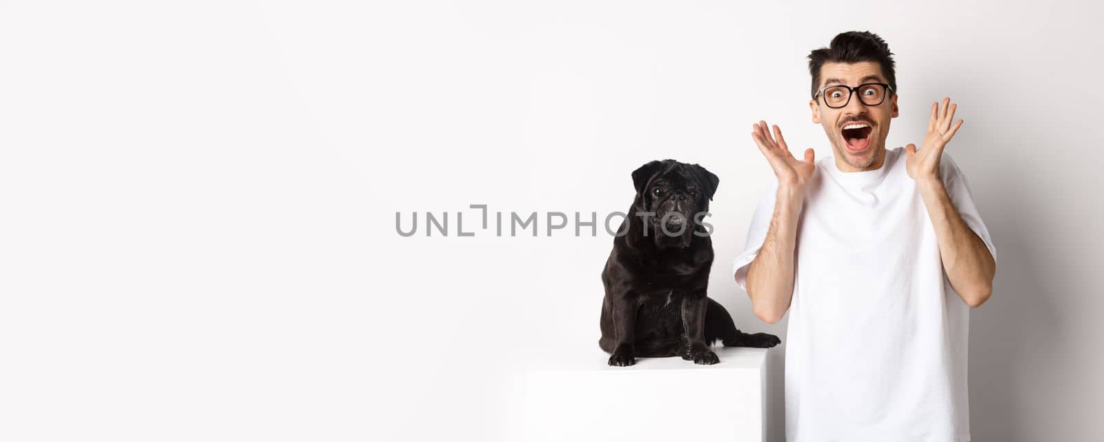 Ecstatic young man looking with excitement and rejoice, standing near cute black pug, staring at camera happy, standing over white background by Benzoix