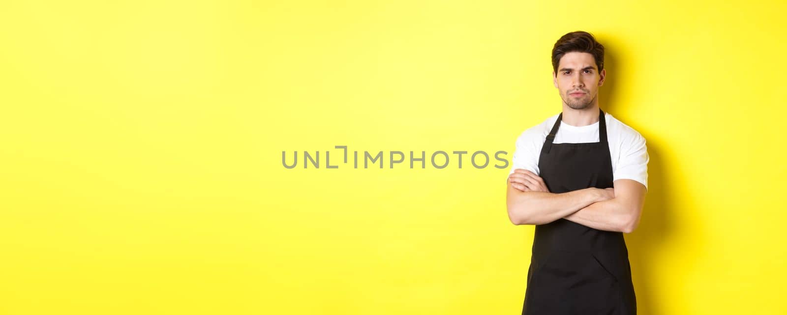 Suspicious barista squinting, cross arms on chest and looking at something with disbelief, standing over yellow background by Benzoix