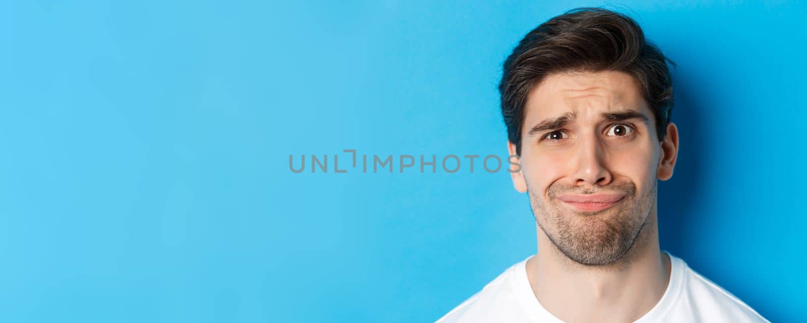 Head shot of skeptical and disappointed man, grimacing complicated, standing against blue background.