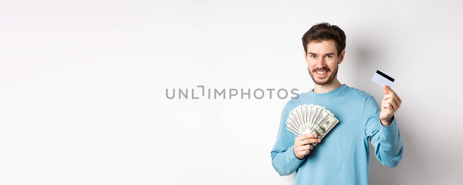Handsome young man smiling and offering payment in cash and contactless, showing money with plastic credit card, standing on white background.