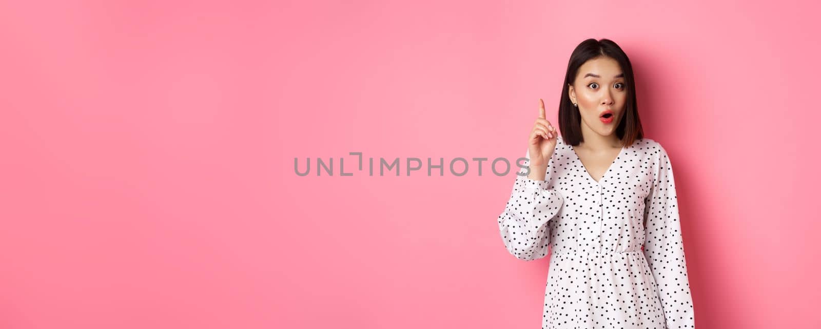 Excited asian girl having an idea, saying suggestion, raising finger and talking, standing over pink background by Benzoix