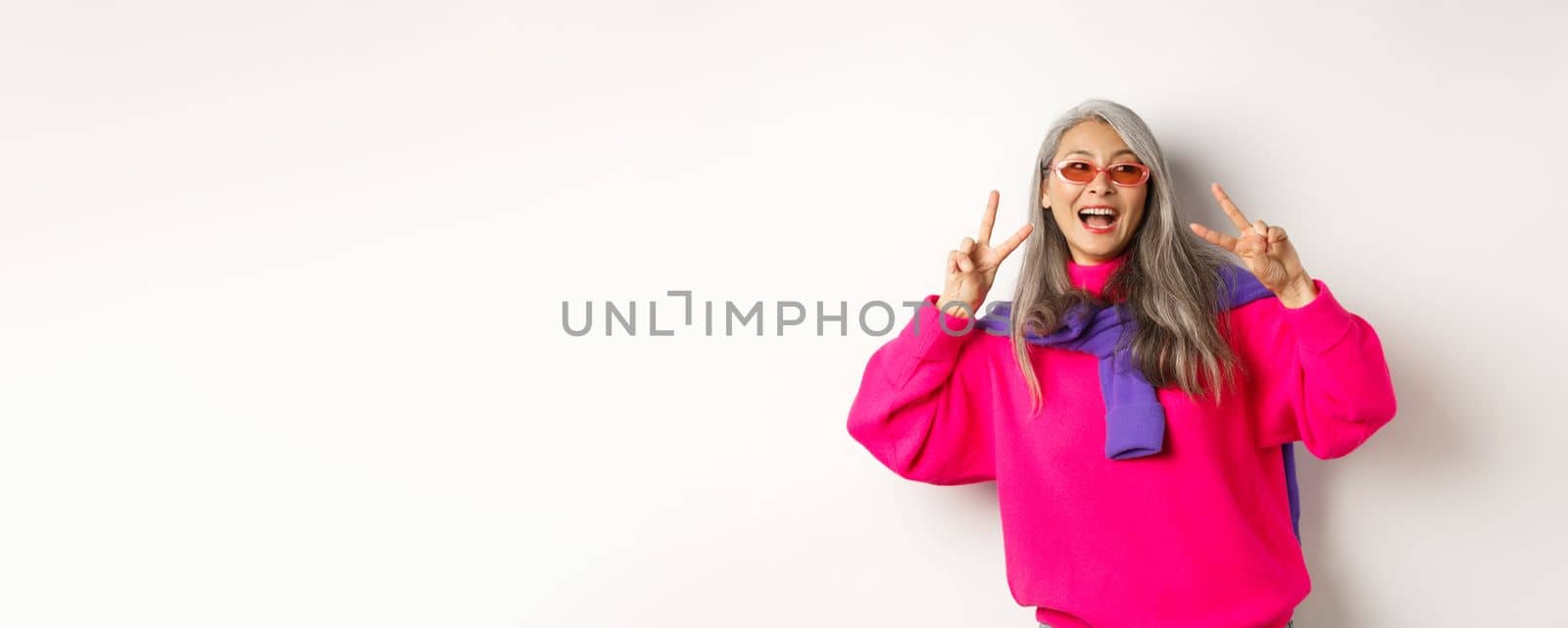 Fashion and beauty concept. Image of stylish asian senior woman in sunglasses smiling, showing peace signs and looking happy, standing over white background.