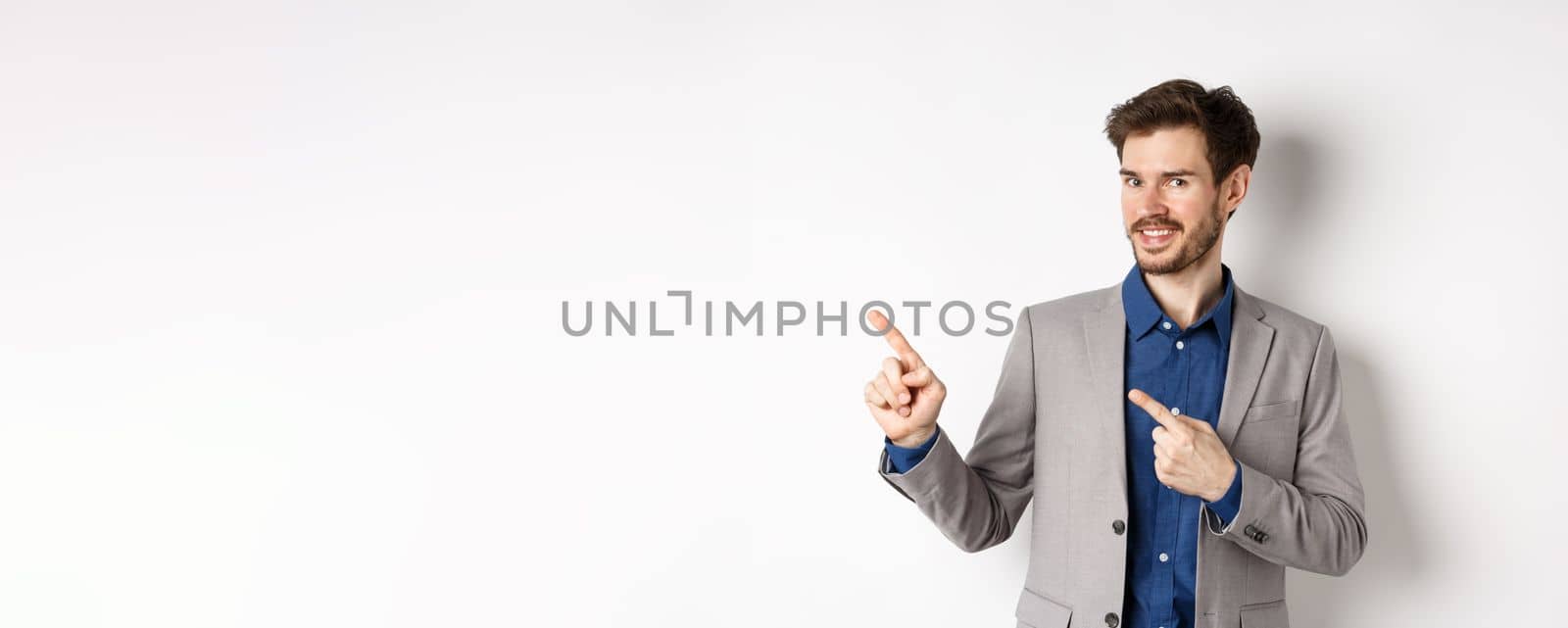 Handsome bearded businessman in grey suit pointing fingers left at logo, inviting check out advertisement, standing on white background.