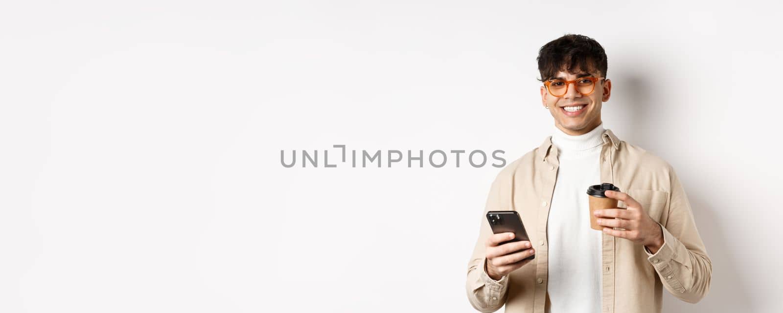 Natural handsome man in glasses drinking coffee from paper cup and using cellphone, smiling satisfied at camera, white background.