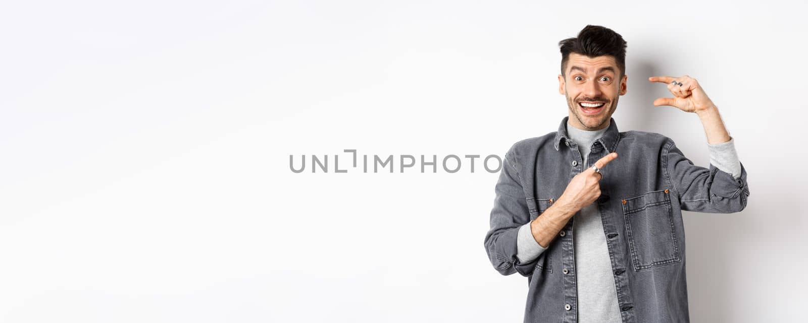 Excited man showing small size, pointing finger at little thing and smiling, standing against white background.
