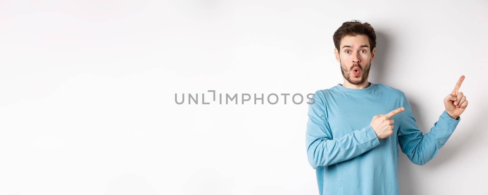 Wow look here. Impressed young man pointing fingers right and showing logo, standing amazed on white background by Benzoix