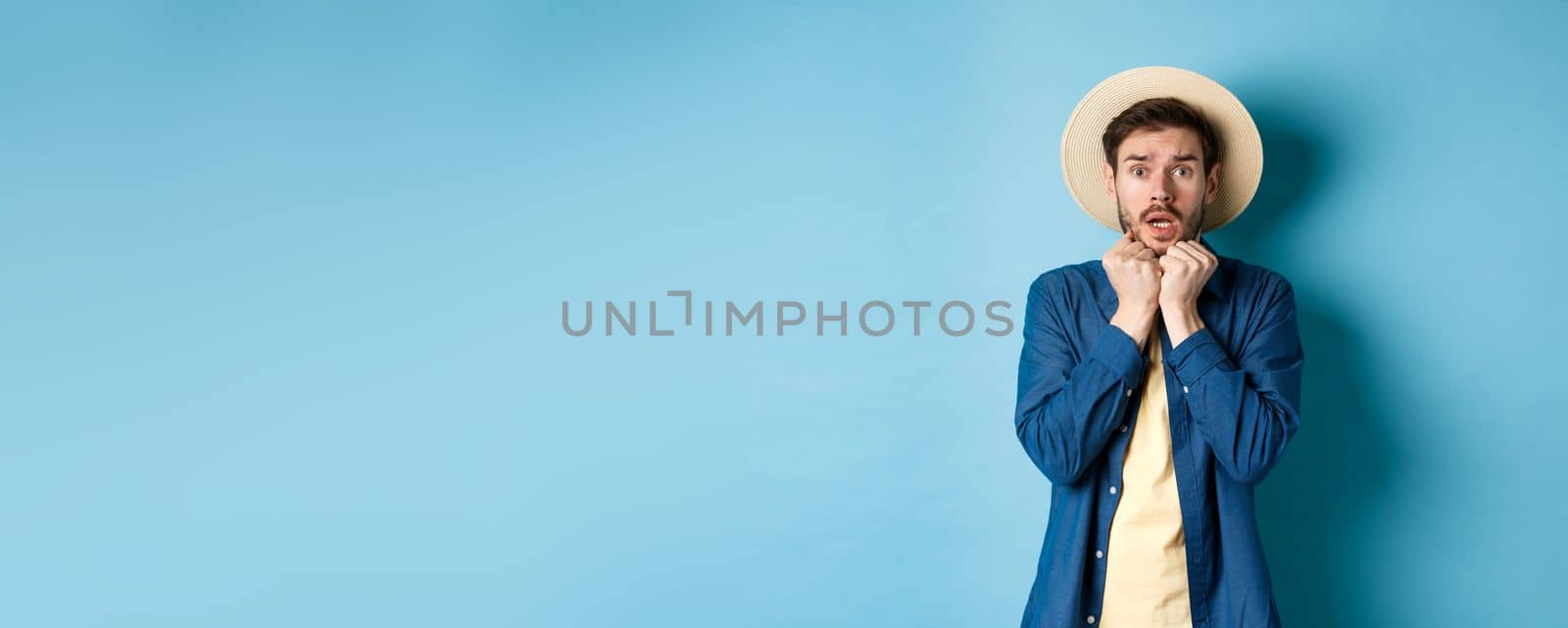 Scared young tourist trembling from fear, looking at something scary on summer vacation, wearing straw hat, standing on blue background.