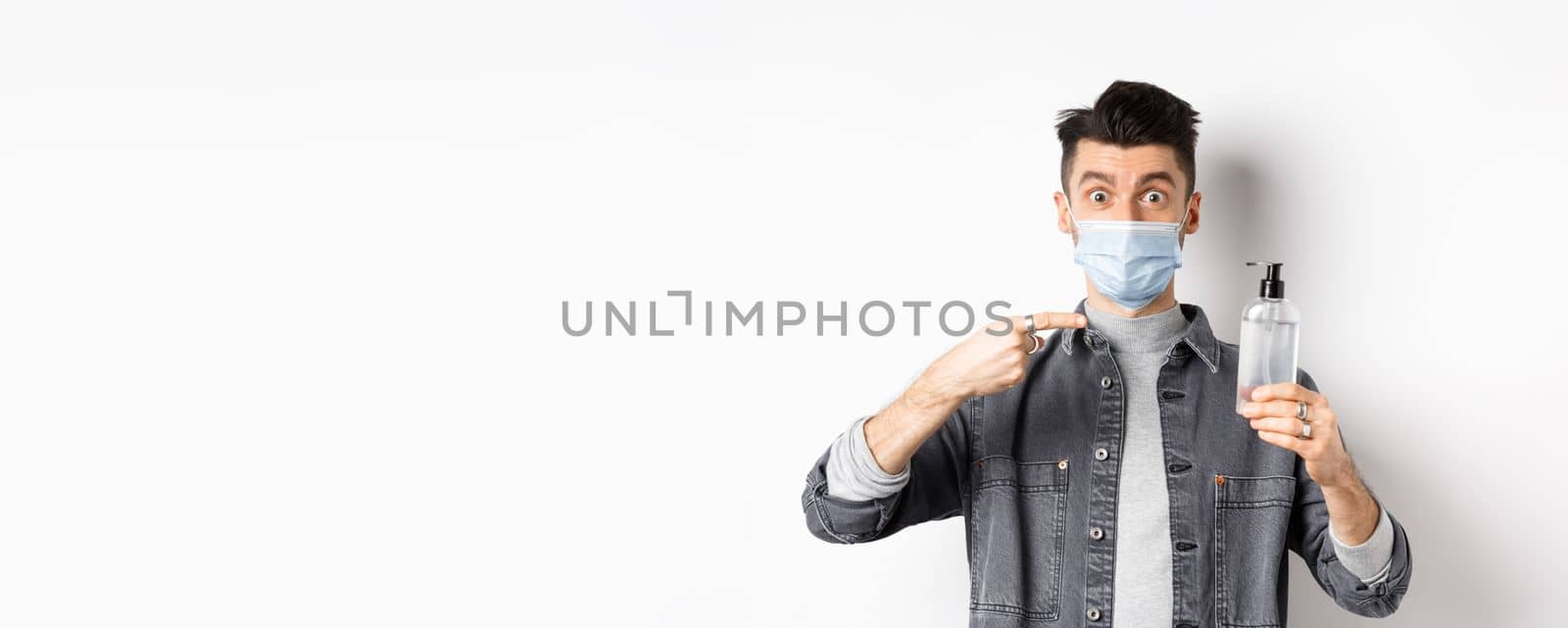 Health, covid and pandemic concept. Stylish guy in medical mask pointing at bottle with hand sanitizer, showing antiseptic, standing on white background.