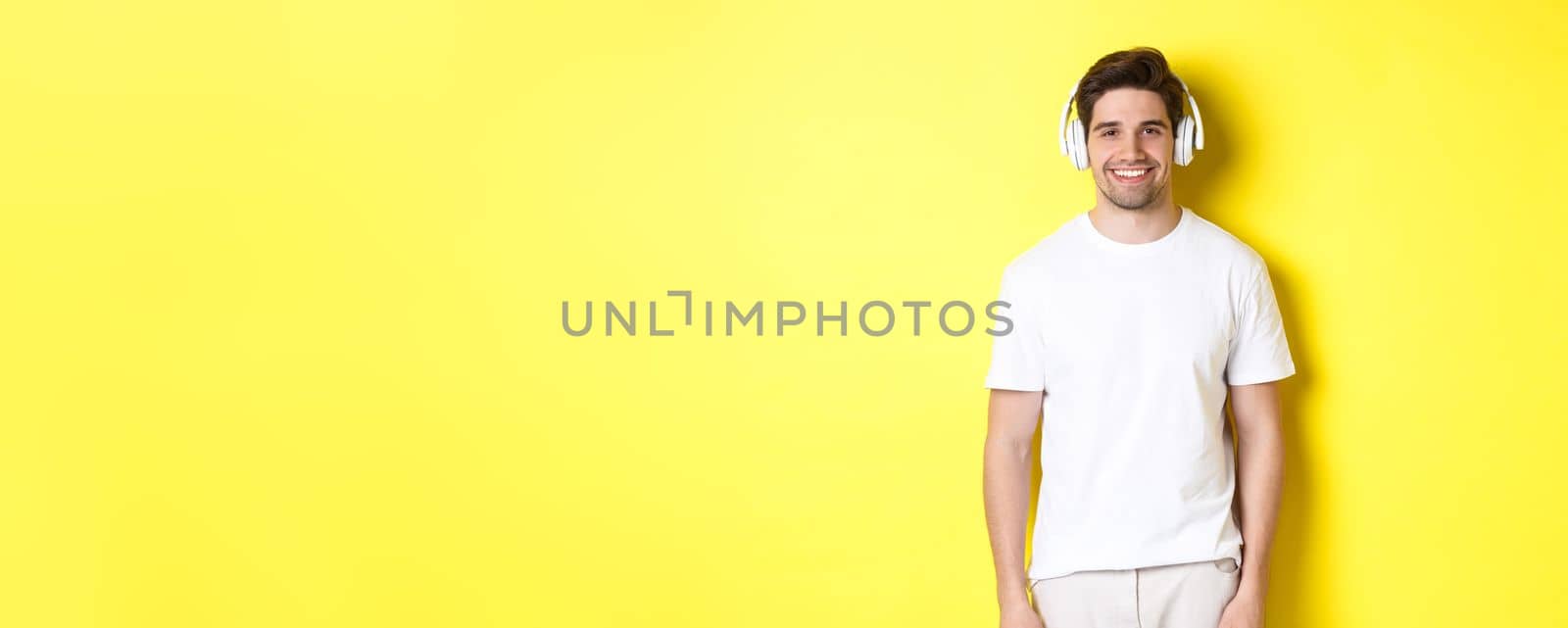 Young handsome man listening music in headphones, wearing earphones and smiling, standing over yellow background.