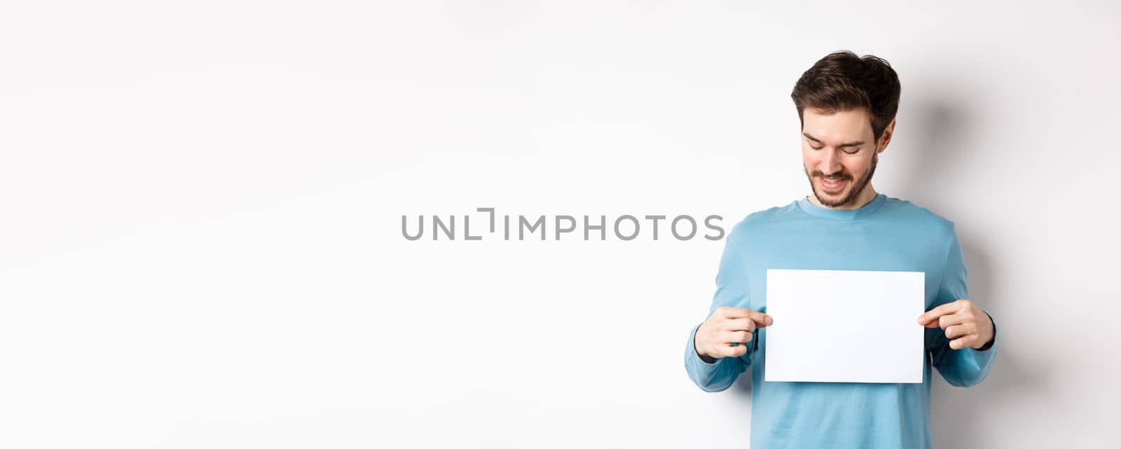 Young caucasian man looking at empty paper, reading logo, standing on white background by Benzoix