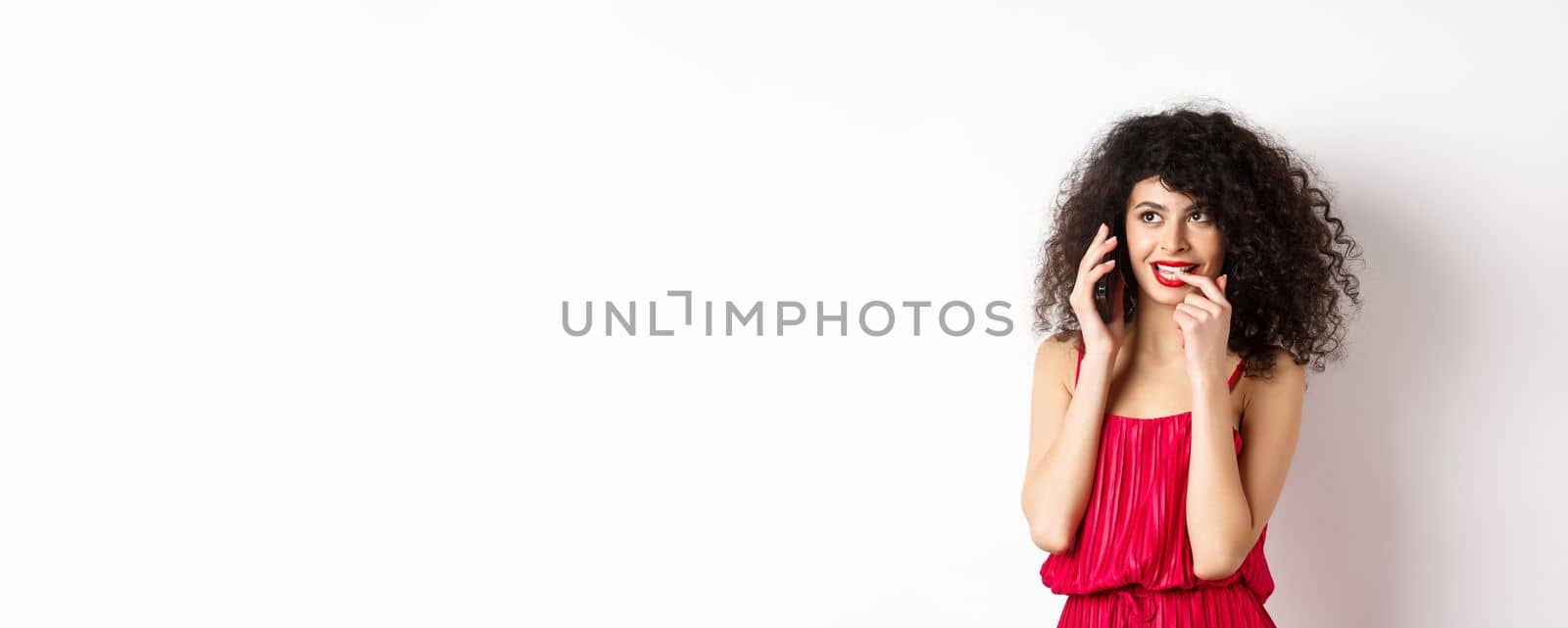 Silly fashionable woman in red dress and lips, biting fingernail duing phone call, thinking on mobile conversation, standing on white background by Benzoix