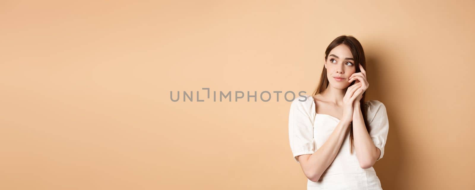 Dreamy and romantic girl looking aside with thoughtful gaze, touching clean gentle facial skin, standing against beige background.