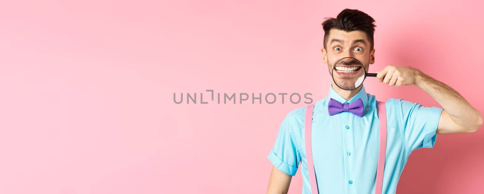 Funny caucasian guy in bow-tie showing his white smile teeth with magnifying glass, looking cheerful at camera, standing on pink background.