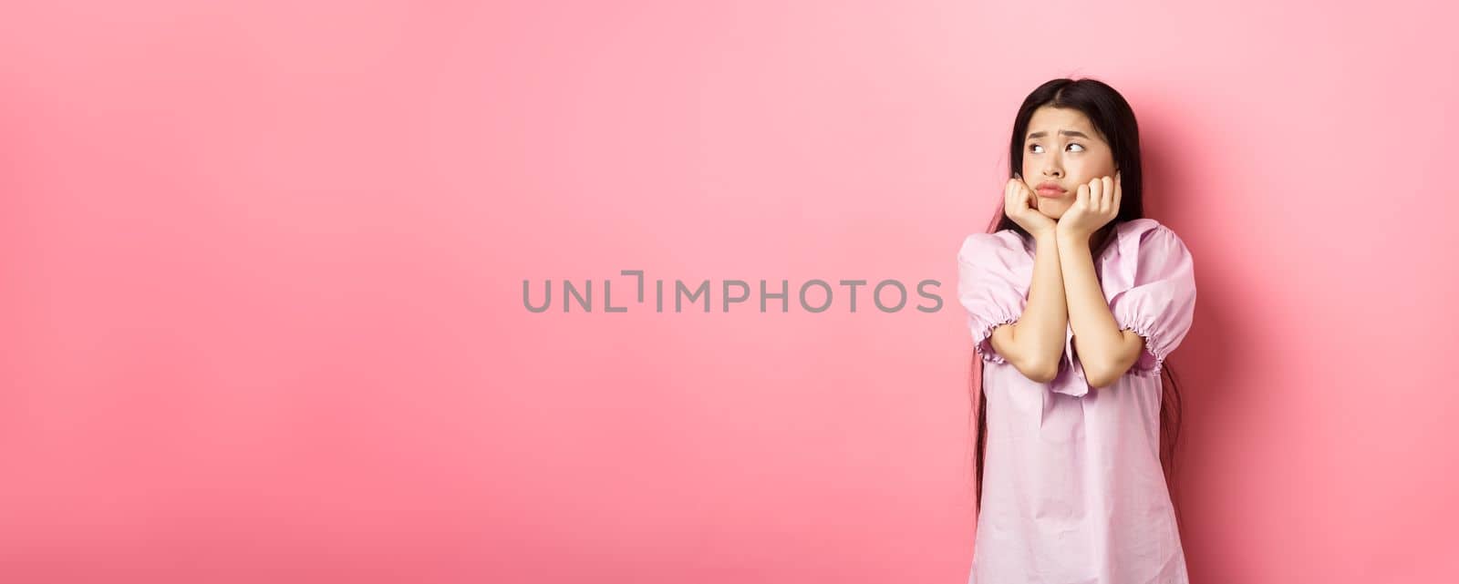 Sad and lonely asian girl look left and frowning, standing distressed against pink background by Benzoix