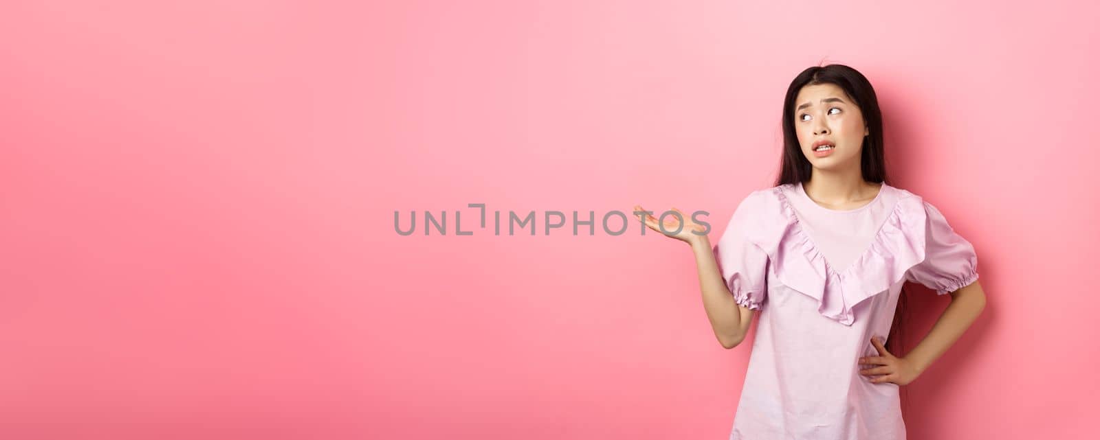 Ignorant asian woman look away and shrugging careless, raising hand up so what gesture, standing unbothered on pink background.