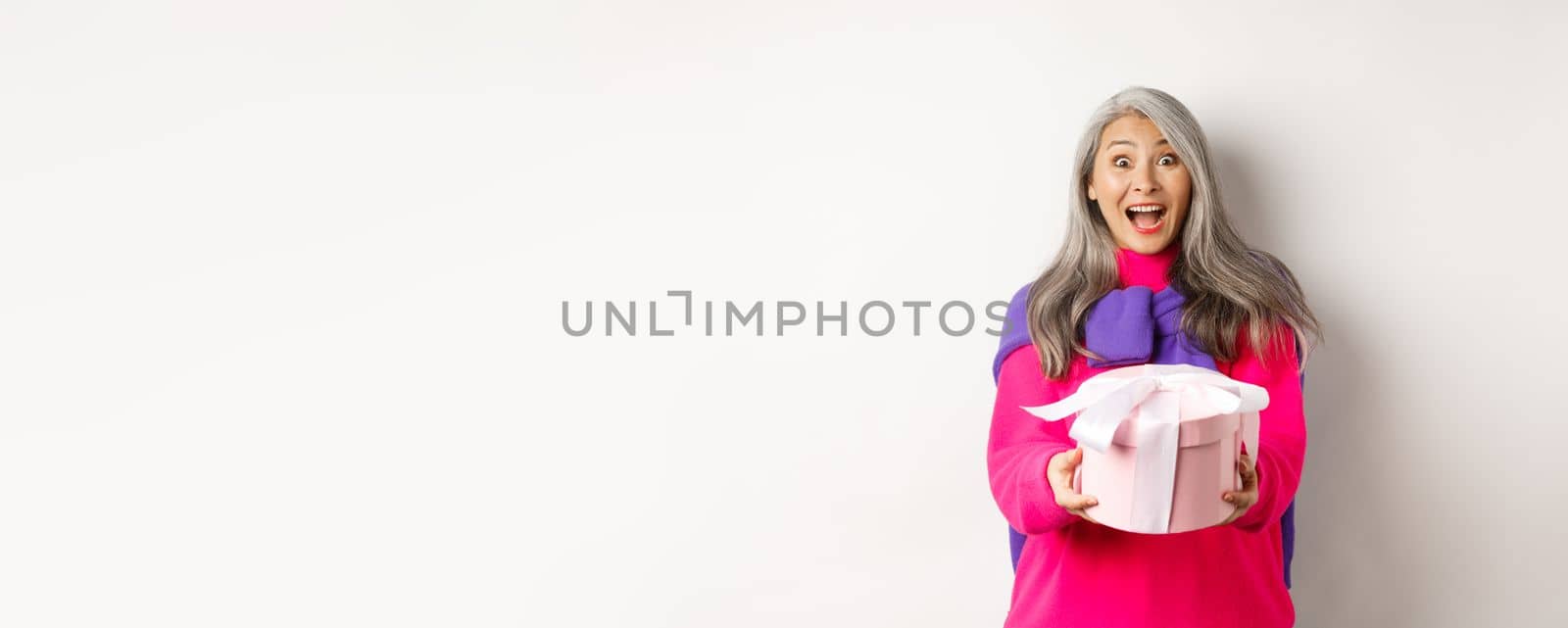 Surprised senior asian woman receiving valentines day gift and smiling amazed, holding present box, standing over white background.