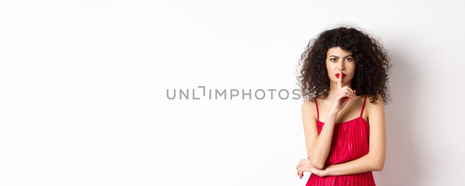 Angry elegant woman in red dress hushing and frowning, tell to be quiet, asking for silence, standing over white background.