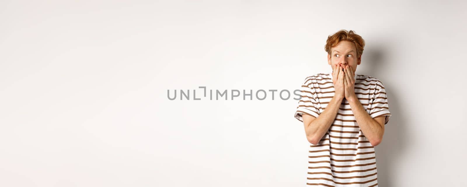 Shocked redhead guy gossiping, giggle in hands and looking left impressed, standing over white background by Benzoix