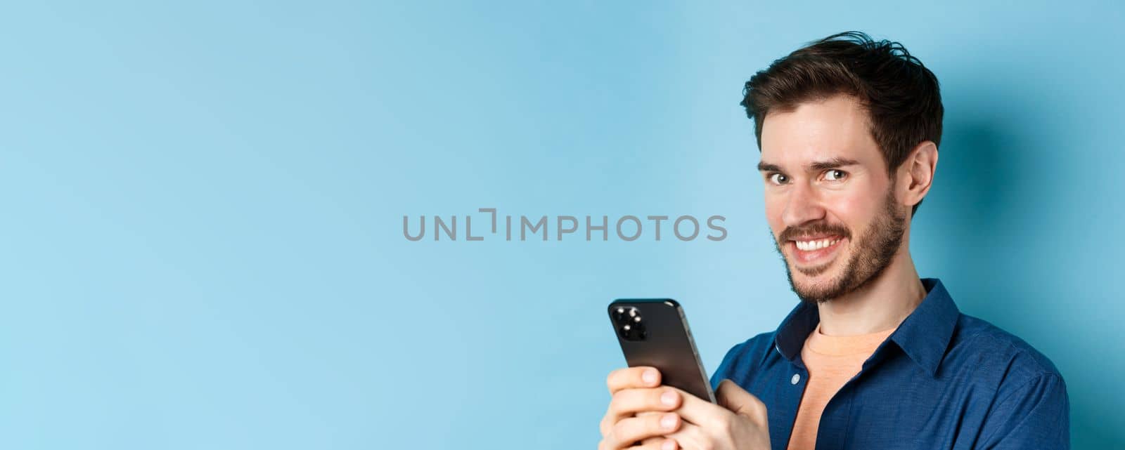 Close-up of cheerful bearded man using smartphone, smiling confident at camera, standing on blue background. Copy space