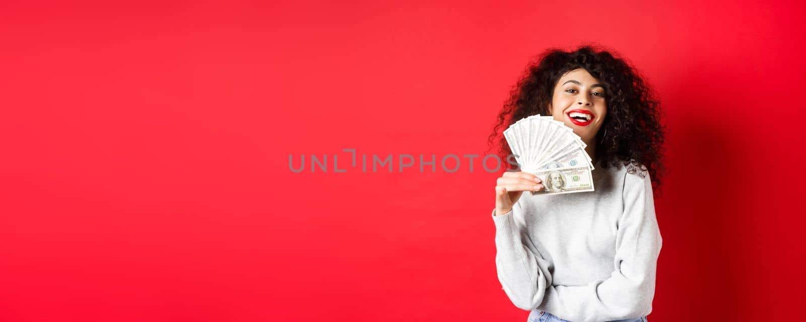 Successful young woman waiving dollar bills and smiling pleased. Rich girl showing money, standing over red background.
