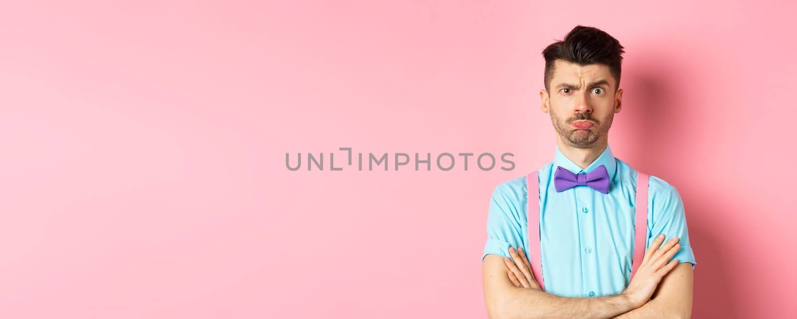 Silly young guy with moustache, sulking and feeling timid, cross arms on chest, being offended at you, standing insulted on pink background by Benzoix