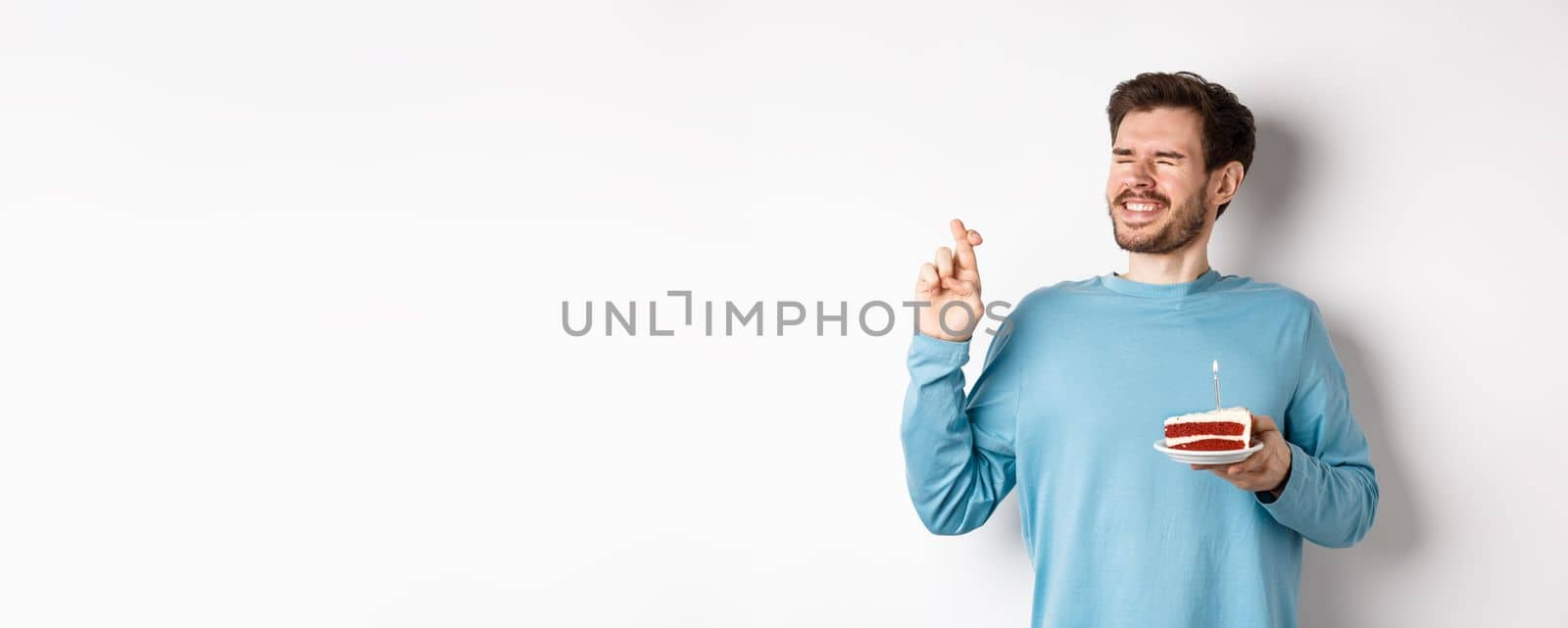 Celebration and holidays concept. Young man celebrating birthday, cross fingers for good luck, making wish on bday cake with lit candle, standing over white background.