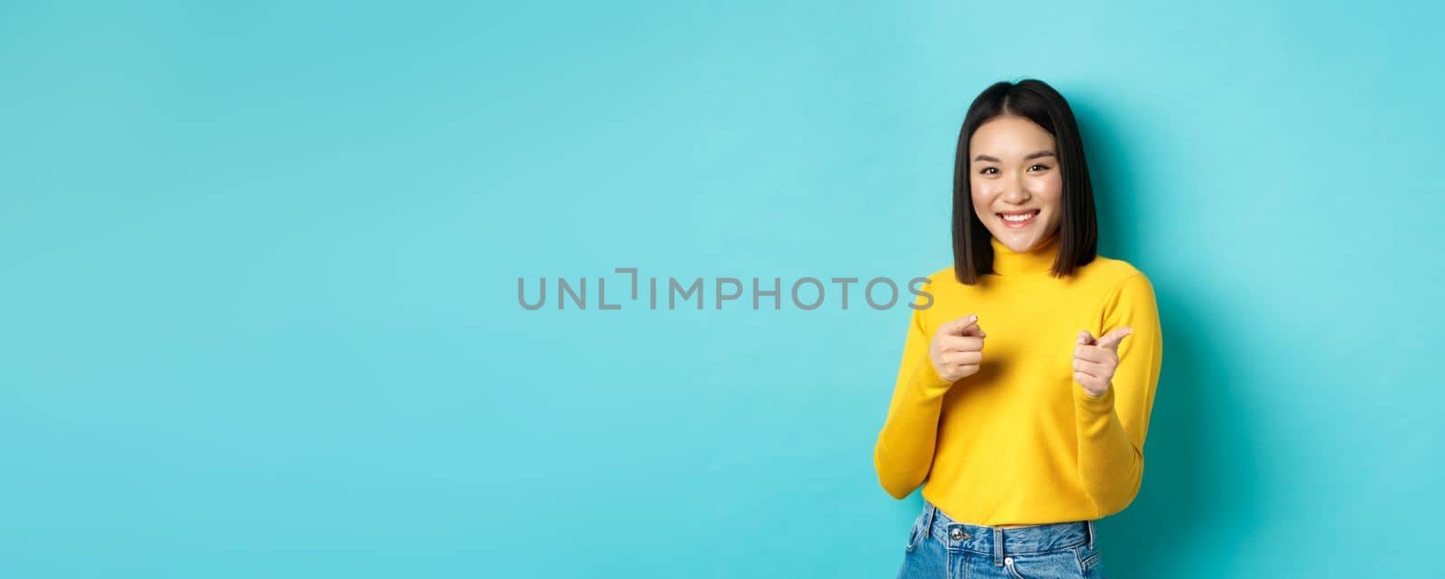 Smiling young asian woman pointing fingers at camera, choosing you, inviting to event, standing happy against blue background by Benzoix
