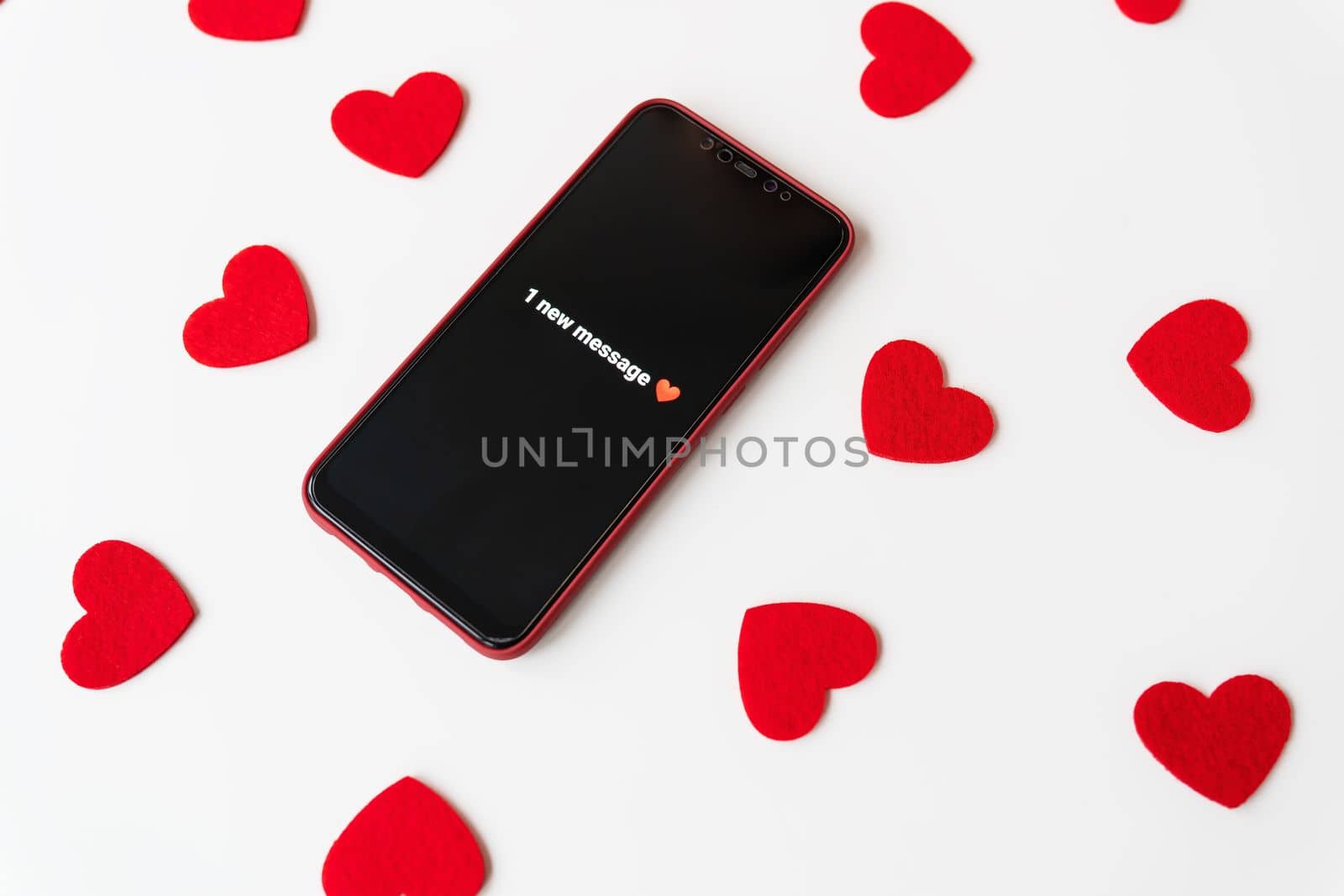 A smartphone with an activated screen with an SMS message in the form of a heart lies on a white table against a background of small red hearts. Business technology and social lifestyle concept. Valentine's Day is February 14