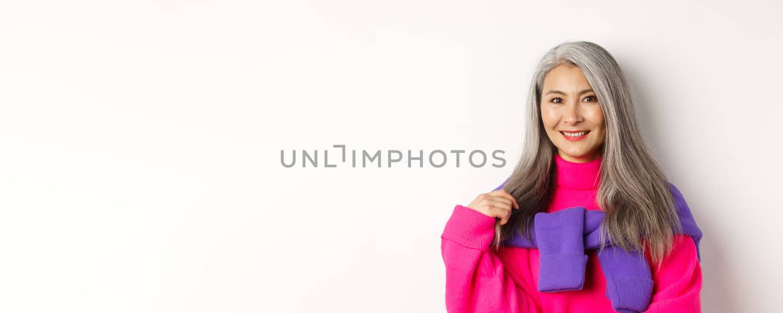 Close-up of beautiful asian senior woman in trendy pink sweater, smiling cheerful at camera, standing over white background.