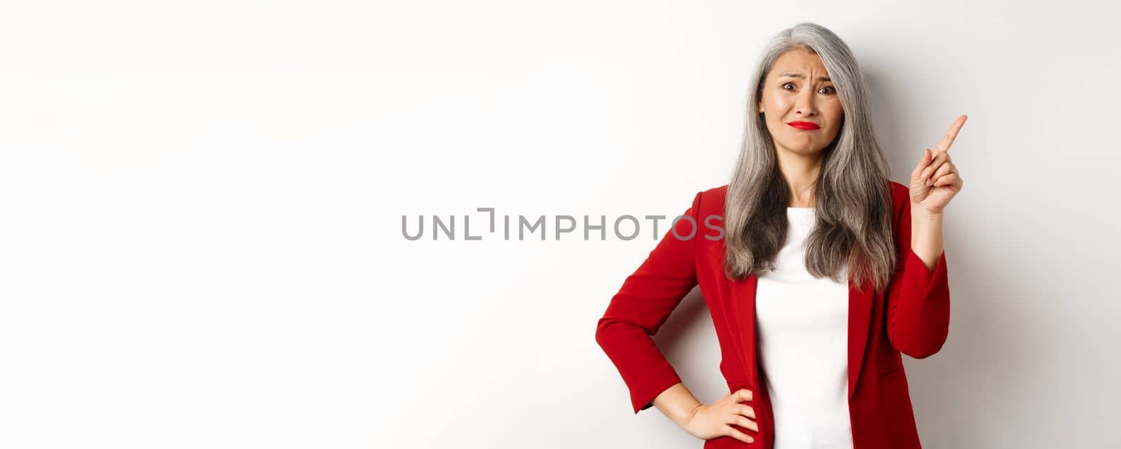 Disappointed and skeptical asian female entrepreneur pointing finger upper right corner, grimacing and frowning upset, showing bad promo, white background.