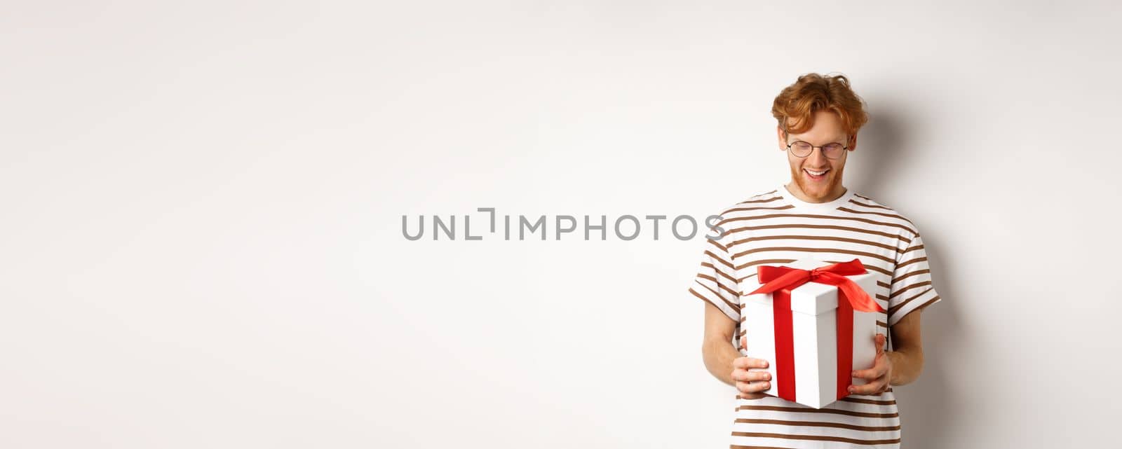 Valentines day and holidays concept. Happy bearded guy holding his gift and smiling, looking at box with present inside, standing over white background by Benzoix