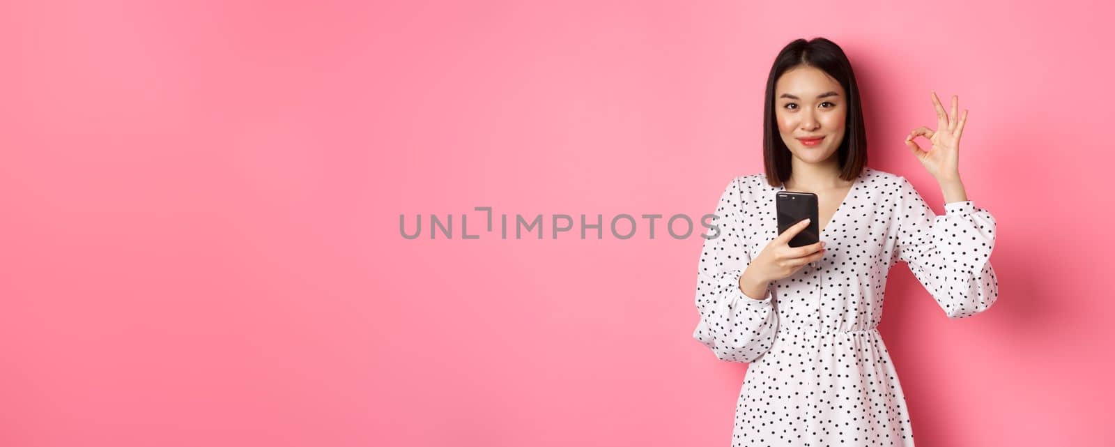 Online shopping and beauty concept. Satisfied asian female customer showing okay, making purchase in internet on smartphone, standing over pink background.