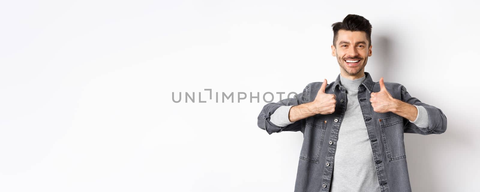 Handsome smiling man showing thumbs-up in approval, praise good work, satisfied with nice choice, recommending something cool, standing on white background.