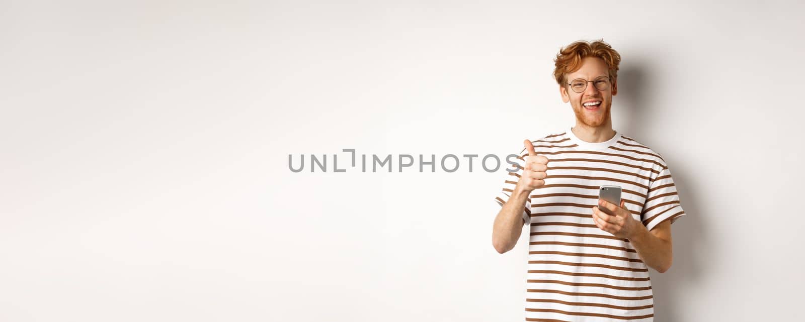 Technology and e-commerce concept. Satisfied male model with red hair, showing thumbs-up and holding smartphone, white background.