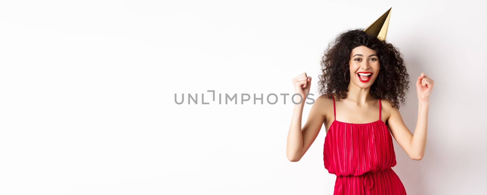 Birthday girl in party hat having fun, dancing in red dress and chanting, standing against white background.