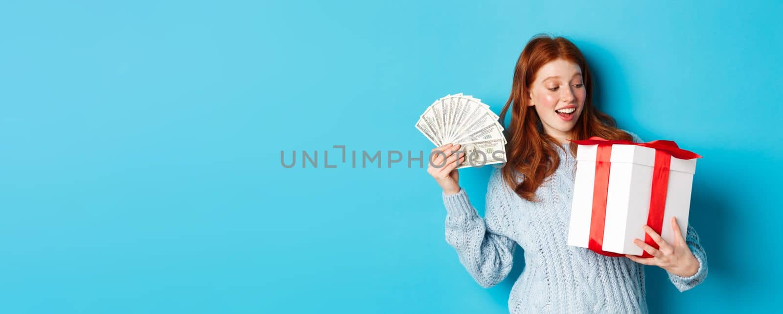 Christmas and shopping concept. Cheerful girl with red hair, holding money and big New Year gift, smiling happy, standing over blue background.