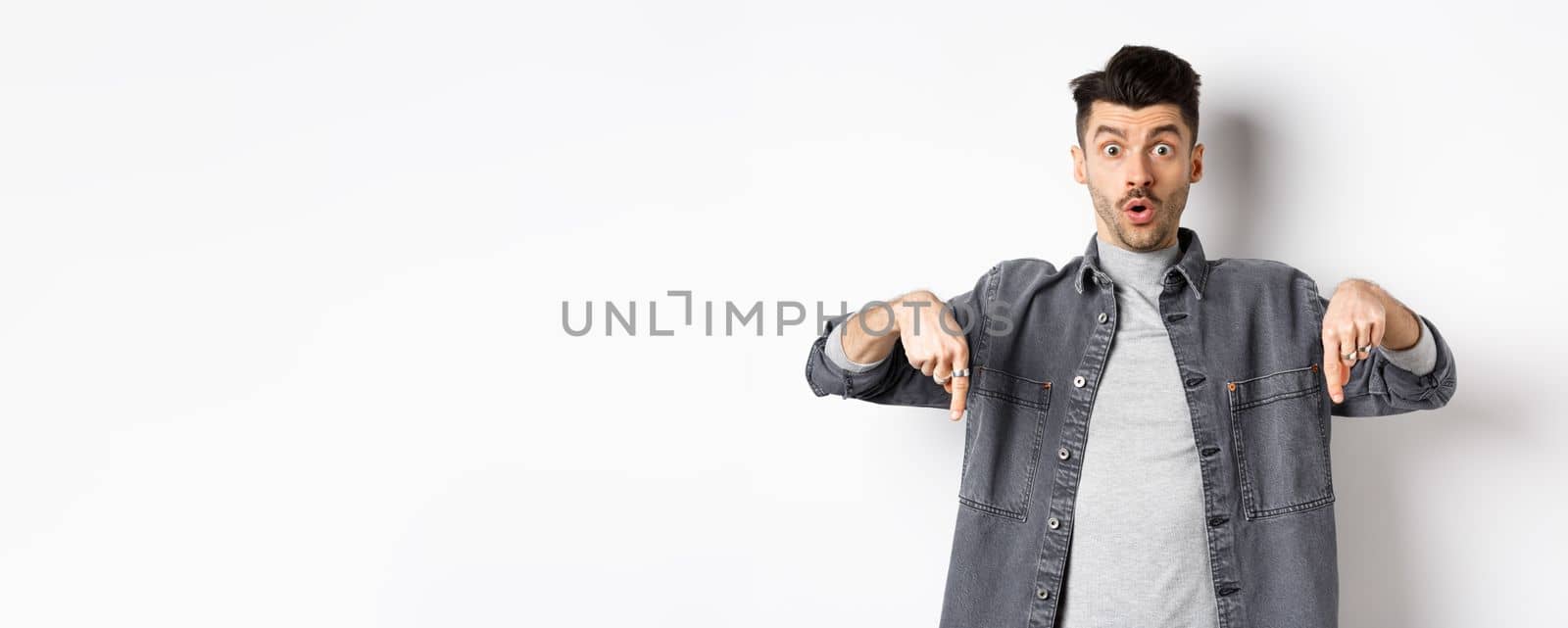 Excited guy say wow, pointing fingers down amazed, showing cool promo offer, standing on white background in denim jacket.